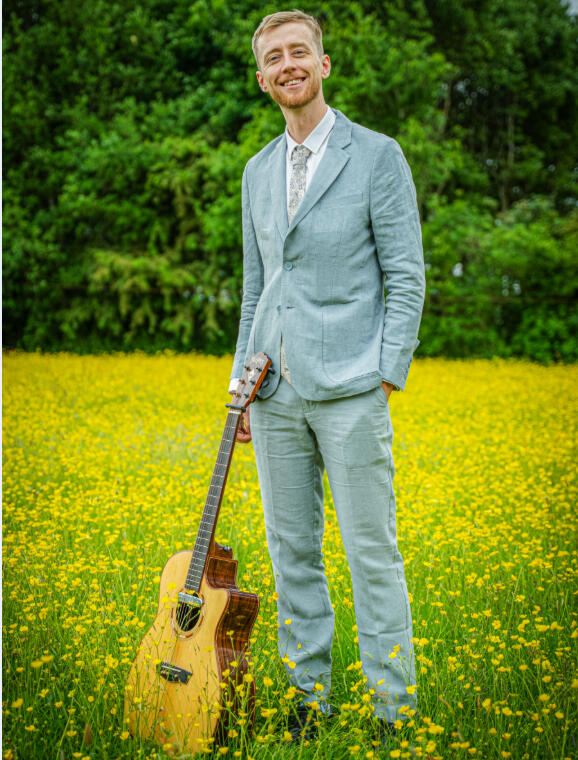 Luke Shaw standing in a field with his guitar wearing a suit. Photo credit: Pete Shaw
