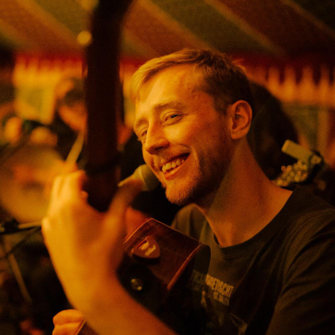 Luke Shaw playing guitar on stage and smiling. Photo credit: Unknown